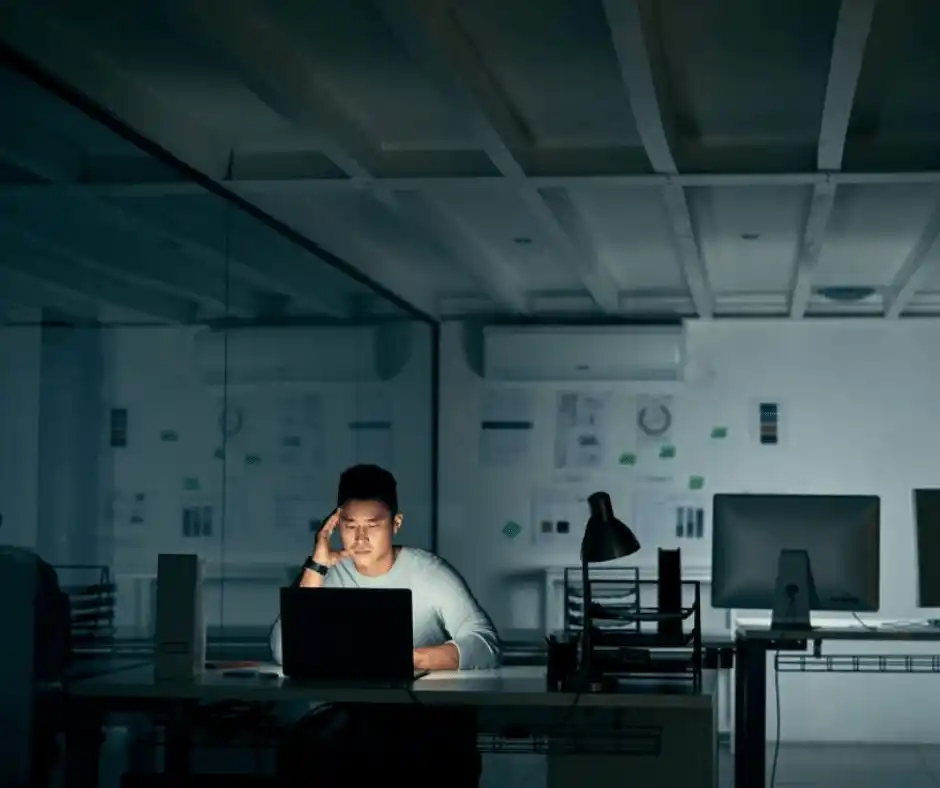 A worker sitting alone in a dimly lit office, looking at a laptop, not given advance notice for changes in working hours by their employer, symbolizing challenges with California Work Schedule Laws.