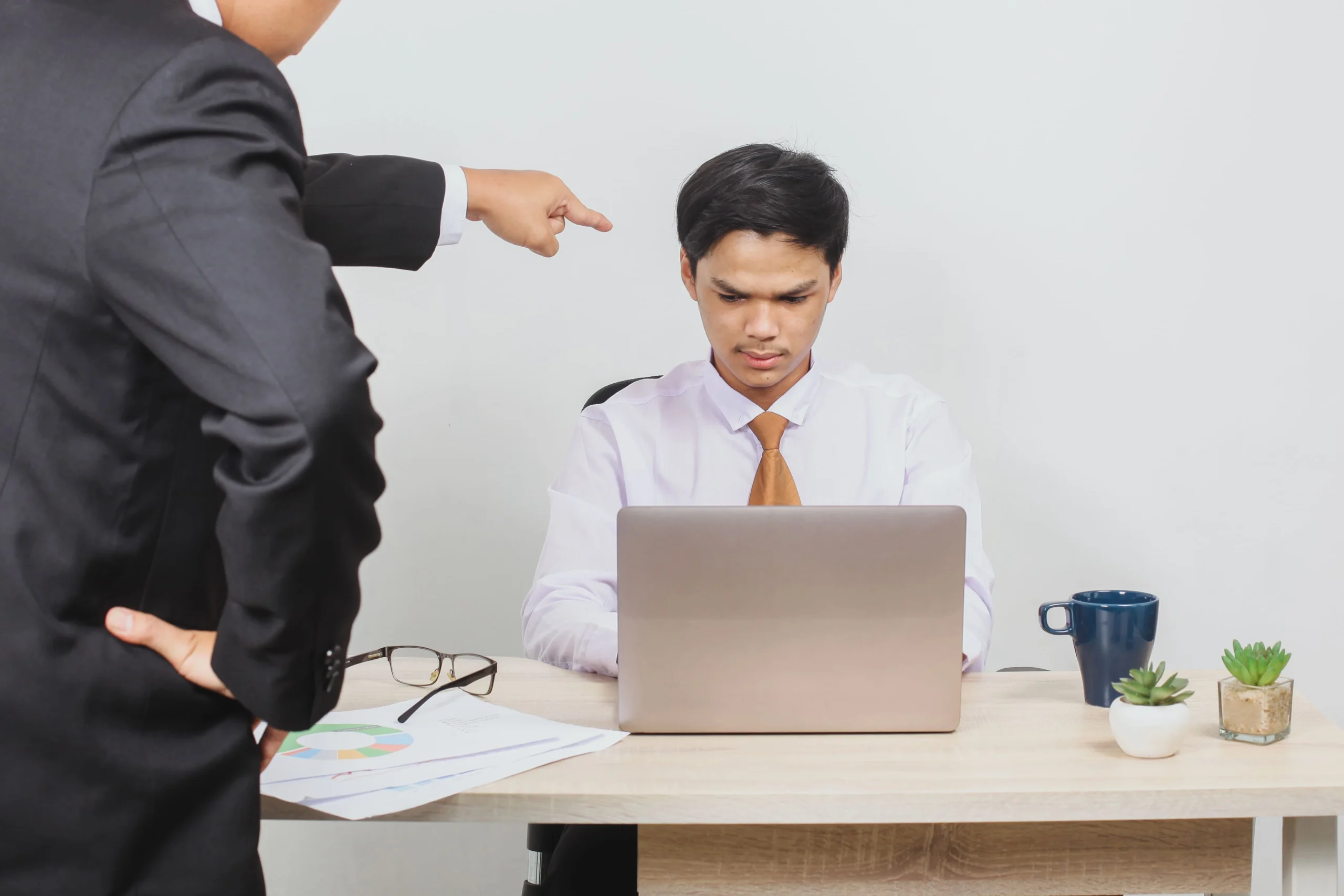 An employee sits at a desk looking stressed while working on a laptop, as a manager in a suit points sternly at him, highlighting workplace conflict.